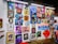A vibrant collection of framed artworks displayed on a gallery wall. The pieces range from colorful portraits to various artistic styles and themes. Each artwork showcases unique characters or figures, while the frames vary in shape and size. The setting appears lively and creative, with another person standing on the right side.