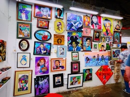 A vibrant collection of framed artworks displayed on a gallery wall. The pieces range from colorful portraits to various artistic styles and themes. Each artwork showcases unique characters or figures, while the frames vary in shape and size. The setting appears lively and creative, with another person standing on the right side.