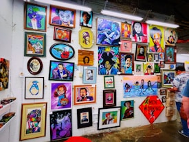 A vibrant collection of framed artworks displayed on a gallery wall. The pieces range from colorful portraits to various artistic styles and themes. Each artwork showcases unique characters or figures, while the frames vary in shape and size. The setting appears lively and creative, with another person standing on the right side.
