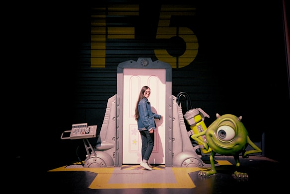 A person stands smiling next to a whimsical door set, accompanied by a large green cartoon character with one eye. The background features a large yellow 'F5' against a dark wall. The setup suggests a theme or amusement park attraction with playful and futuristic elements.