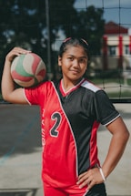 A young athlete stands confidently in a red and black sports uniform with the number 2, holding a volleyball under one arm. The setting appears to be an outdoor volleyball court, indicated by the net behind her.
