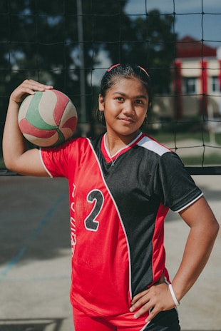 A young athlete stands confidently in a red and black sports uniform with the number 2, holding a volleyball under one arm. The setting appears to be an outdoor volleyball court, indicated by the net behind her.