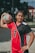 A young athlete stands confidently in a red and black sports uniform with the number 2, holding a volleyball under one arm. The setting appears to be an outdoor volleyball court, indicated by the net behind her.