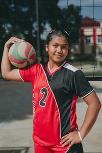 A young athlete stands confidently in a red and black sports uniform with the number 2, holding a volleyball under one arm. The setting appears to be an outdoor volleyball court, indicated by the net behind her.