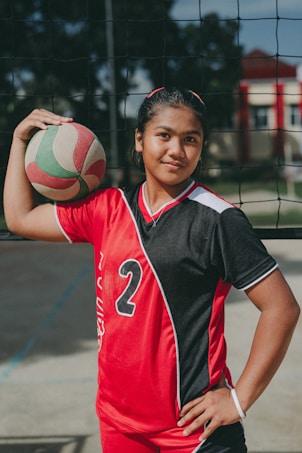 A young athlete stands confidently in a red and black sports uniform with the number 2, holding a volleyball under one arm. The setting appears to be an outdoor volleyball court, indicated by the net behind her.