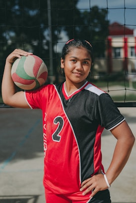 A young athlete stands confidently in a red and black sports uniform with the number 2, holding a volleyball under one arm. The setting appears to be an outdoor volleyball court, indicated by the net behind her.