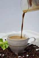 A stream of coffee is being poured into a white cup surrounded by coffee beans. A green leaf adds a touch of color to the composition, placed near the cup. The background is a plain light color, enhancing the focus on the coffee and the beans.