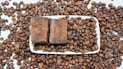 A large number of roasted coffee beans scattered across a white surface, with a rectangular white dish at the center containing two pieces of brown, rustic-looking soap. The coffee beans are evenly spread both on and around the dish.