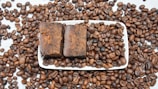 A large number of roasted coffee beans scattered across a white surface, with a rectangular white dish at the center containing two pieces of brown, rustic-looking soap. The coffee beans are evenly spread both on and around the dish.
