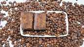 A large number of roasted coffee beans scattered across a white surface, with a rectangular white dish at the center containing two pieces of brown, rustic-looking soap. The coffee beans are evenly spread both on and around the dish.