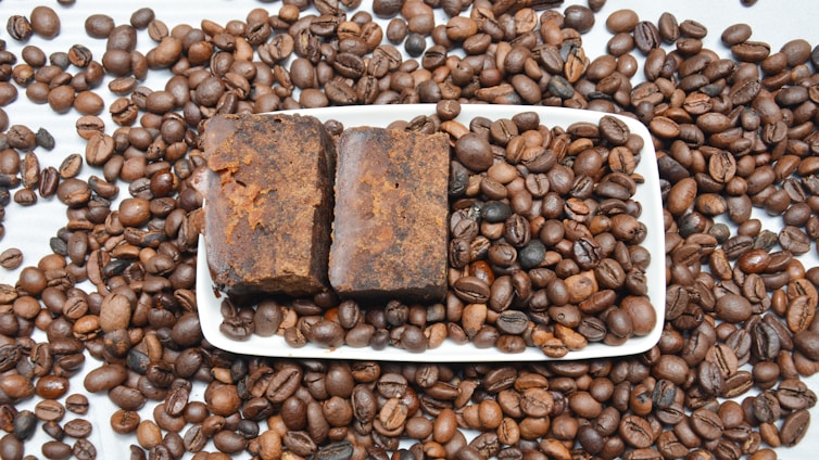 A large number of roasted coffee beans scattered across a white surface, with a rectangular white dish at the center containing two pieces of brown, rustic-looking soap. The coffee beans are evenly spread both on and around the dish.