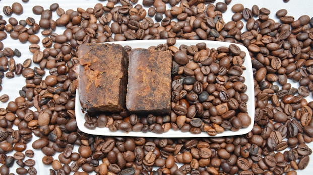 A large number of roasted coffee beans scattered across a white surface, with a rectangular white dish at the center containing two pieces of brown, rustic-looking soap. The coffee beans are evenly spread both on and around the dish.