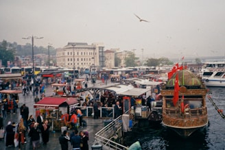 A bustling waterfront scene with numerous people walking around an area filled with food stalls and market vendors. A large, ornately decorated boat with Turkish flags is docked in the foreground. Buildings and a bridge are visible in the background, and several buses and cars are parked nearby. The sky is overcast, and seagulls are flying overhead.