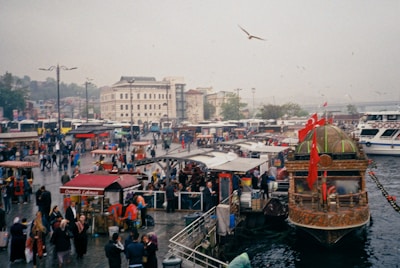 A bustling waterfront scene with numerous people walking around an area filled with food stalls and market vendors. A large, ornately decorated boat with Turkish flags is docked in the foreground. Buildings and a bridge are visible in the background, and several buses and cars are parked nearby. The sky is overcast, and seagulls are flying overhead.