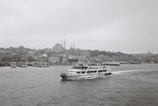 A black and white photograph captures a cityscape featuring numerous buildings along the waterfront. A ferry with the name 'Turyol' prominently visible sails across the water towards the right. The skyline includes several domed structures and minarets, hinting at historical architecture. The overall atmosphere is calm and serene, with the sea slightly rippling under the overcast sky.