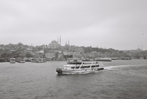 A black and white photograph captures a cityscape featuring numerous buildings along the waterfront. A ferry with the name 'Turyol' prominently visible sails across the water towards the right. The skyline includes several domed structures and minarets, hinting at historical architecture. The overall atmosphere is calm and serene, with the sea slightly rippling under the overcast sky.