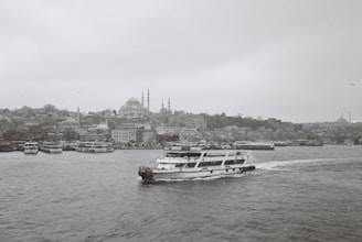 A black and white photograph captures a cityscape featuring numerous buildings along the waterfront. A ferry with the name 'Turyol' prominently visible sails across the water towards the right. The skyline includes several domed structures and minarets, hinting at historical architecture. The overall atmosphere is calm and serene, with the sea slightly rippling under the overcast sky.