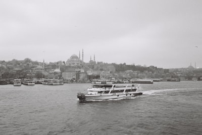 A black and white photograph captures a cityscape featuring numerous buildings along the waterfront. A ferry with the name 'Turyol' prominently visible sails across the water towards the right. The skyline includes several domed structures and minarets, hinting at historical architecture. The overall atmosphere is calm and serene, with the sea slightly rippling under the overcast sky.
