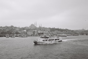 A black and white photograph captures a cityscape featuring numerous buildings along the waterfront. A ferry with the name 'Turyol' prominently visible sails across the water towards the right. The skyline includes several domed structures and minarets, hinting at historical architecture. The overall atmosphere is calm and serene, with the sea slightly rippling under the overcast sky.