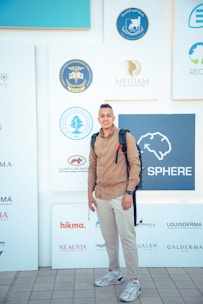 A man is standing in front of a backdrop featuring various logos related to medical and dermatological organizations. He is wearing a brown shirt, light-colored pants, and sneakers, with a backpack on his shoulders.