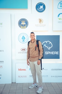 A man is standing in front of a backdrop featuring various logos related to medical and dermatological organizations. He is wearing a brown shirt, light-colored pants, and sneakers, with a backpack on his shoulders.