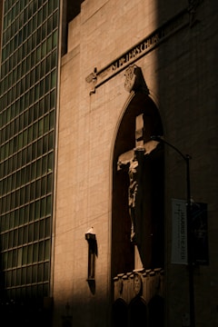 A tall, sunlit building facade features an arched niche with a crucifix displaying a prominent sculpture of Jesus. The architecture is classical, with detailed engravings and inscriptions. Adjacent, the modern grid design of a glass and steel skyscraper casts a shadow.