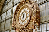 An ornate, large clock with Roman numerals and intricate gold detailing is mounted on an interior wall featuring metal framework and large glass panels. The clock's elaborate design includes decorative elements and appears to be a historical or classical style.