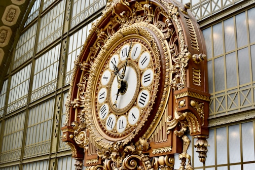 An ornate, large clock with Roman numerals and intricate gold detailing is mounted on an interior wall featuring metal framework and large glass panels. The clock's elaborate design includes decorative elements and appears to be a historical or classical style.