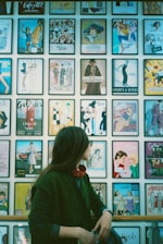 A person gazes at a wall filled with framed vintage magazine covers featuring various fashion designs and illustrations. The individual is wearing a dark green coat and a red scarf.