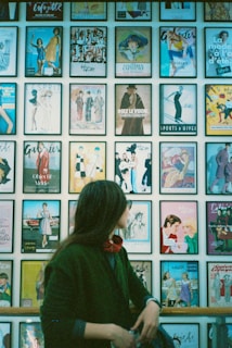 A person gazes at a wall filled with framed vintage magazine covers featuring various fashion designs and illustrations. The individual is wearing a dark green coat and a red scarf.