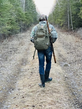 A person is walking down a forest path, carrying a rifle slung over their shoulder and wearing camouflage clothing, rubber boots, and a backpack. The tall trees on either side create a tunnel-like effect and the ground is covered with dry leaves and grass.