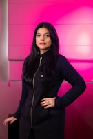 A woman with long dark hair stands confidently against a vibrant pink background. She is wearing a black outfit with a zipper running down the front and a small crown emblem near the chest. Her makeup is neatly done, and she has red nail polish on her fingers.