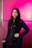 A woman with long dark hair stands confidently against a vibrant pink background. She is wearing a black outfit with a zipper running down the front and a small crown emblem near the chest. Her makeup is neatly done, and she has red nail polish on her fingers.