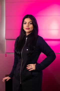 A woman with long dark hair stands confidently against a vibrant pink background. She is wearing a black outfit with a zipper running down the front and a small crown emblem near the chest. Her makeup is neatly done, and she has red nail polish on her fingers.