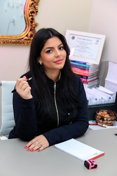 A woman with long dark hair is seated at a desk holding a pen. She is wearing a dark zip-up jacket and has red painted nails. Behind her, certificates and stacks of colorful books are visible. A gold-framed mirror is partially visible on the wall.