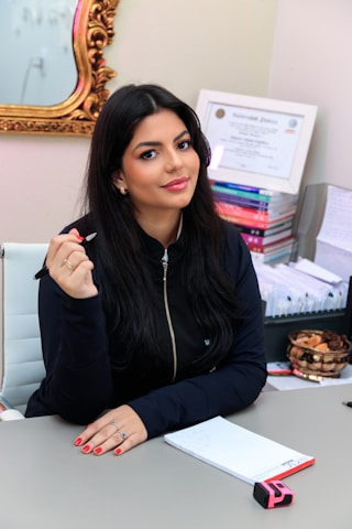 A woman with long dark hair is seated at a desk holding a pen. She is wearing a dark zip-up jacket and has red painted nails. Behind her, certificates and stacks of colorful books are visible. A gold-framed mirror is partially visible on the wall.