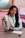 A person with long dark hair is seated at a desk wearing a white blazer with black trim, holding a pencil. They are positioned in front of a mirror with an ornate gold frame. To the side, there is a stack of books and a certificate on the wall. A notepad and a pink and black object are on the desk.