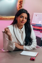A person with long dark hair is seated at a desk wearing a white blazer with black trim, holding a pencil. They are positioned in front of a mirror with an ornate gold frame. To the side, there is a stack of books and a certificate on the wall. A notepad and a pink and black object are on the desk.