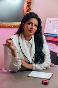A person with long dark hair is seated at a desk wearing a white blazer with black trim, holding a pencil. They are positioned in front of a mirror with an ornate gold frame. To the side, there is a stack of books and a certificate on the wall. A notepad and a pink and black object are on the desk.