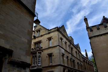 Historic stone buildings with intricate architectural details are seen against a backdrop of a partly cloudy blue sky. The structures feature large windows and detailed ornamentation, suggesting an old, possibly academic or religious setting.