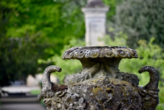 A weathered stone urn, covered in moss and lichen, stands prominently against a blurred background of greenery. The urn's ornate design features intricate carvings and two handles on either side.