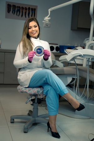 A person is sitting on a dentist's chair holding a round plaque, wearing a white coat and pink gloves in what appears to be a dental office. The room contains dental equipment and artwork depicting teeth.