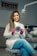 A woman wearing a white lab coat and blue pants is seated on a cushioned chair in a dental office. She is holding a circular object with the word 'Elastomerics' on it. The setting includes dental equipment and a light fixture above her.