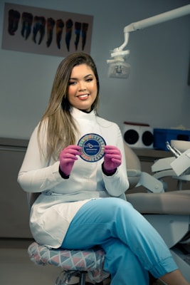 A woman wearing a white lab coat and blue pants is seated on a cushioned chair in a dental office. She is holding a circular object with the word 'Elastomerics' on it. The setting includes dental equipment and a light fixture above her.