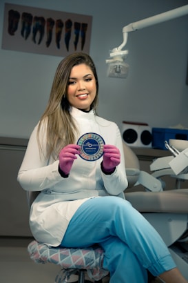 A woman wearing a white lab coat and blue pants is seated on a cushioned chair in a dental office. She is holding a circular object with the word 'Elastomerics' on it. The setting includes dental equipment and a light fixture above her.