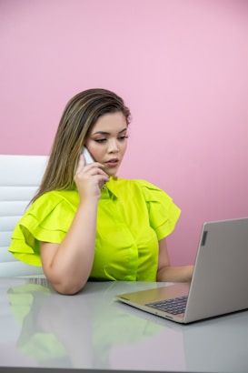 A woman is sitting at a desk, talking on a smartphone while looking at a laptop. She is wearing a bright green blouse, and the background wall is pink. The overall setting seems office-like, with a modern and minimalistic design.