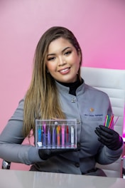 A woman with long hair is wearing a gray uniform and black gloves. She is holding a transparent box containing colorful dental tools, as well as additional colorful dental tools in her other hand. The background is a pink wall and she is sitting on a white chair.