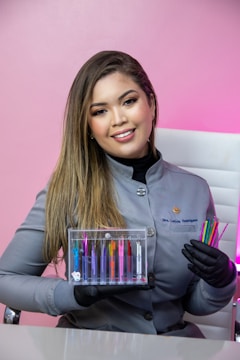 A woman with long hair is wearing a gray uniform and black gloves. She is holding a transparent box containing colorful dental tools, as well as additional colorful dental tools in her other hand. The background is a pink wall and she is sitting on a white chair.