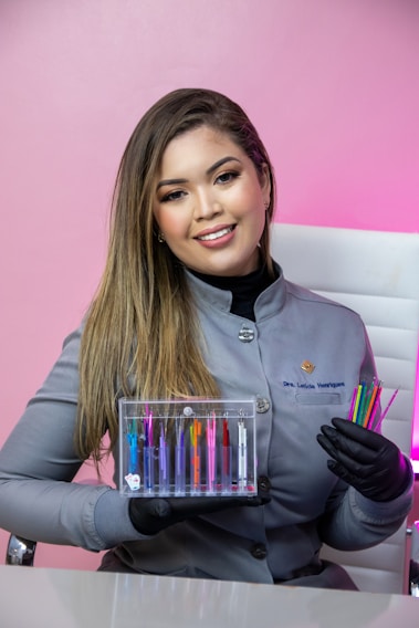 A woman with long hair is wearing a gray uniform and black gloves. She is holding a transparent box containing colorful dental tools, as well as additional colorful dental tools in her other hand. The background is a pink wall and she is sitting on a white chair.