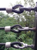 Metal cable clamps securing thick black ropes to a wooden post are prominently featured, against a backdrop of lush green foliage. The metal components have a shiny appearance with droplets of water indicating recent rain.